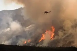 A helicopter hovering over a fierce wildfire, with flames engulfing the hillside and a massive cloud of smoke billowing into the sky.