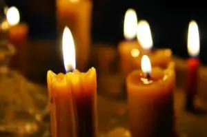 A close-up of several lit wax candles glowing warmly in a dimly lit environment, with the focus on the flame of the closest candle as it casts a soft light on the surroundings.