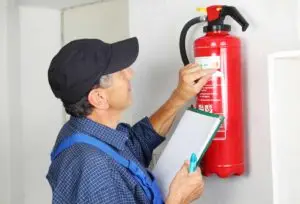 A technician in a cap and plaid shirt inspects a red fire extinguisher mounted on the wall, holding a clipboard for recording details.