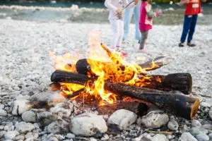 A cozy bonfire crackles with warmth on a bed of rocks, while in the background, people enjoy a leisurely summer gathering.