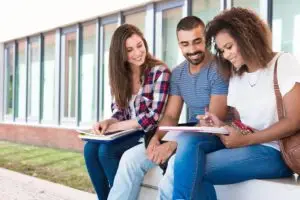 Three university students smiling and studying together outdoors on a sunny day.