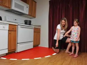 A mother appears to be teaching her young daughter about safety by illustrating the concept of a "no-go" zone around the hot stove with a semicircle of white paper on the kitchen floor.