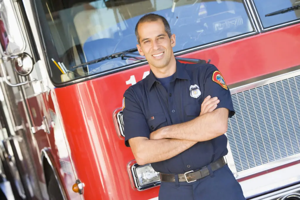 A confident firefighter stands with arms crossed in front of a firetruck, ready to respond to emergencies.