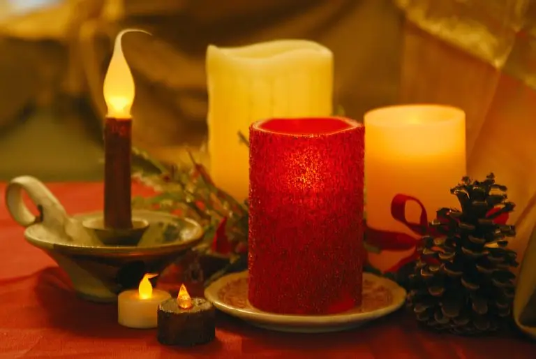 A warm and cozy holiday setting with lit candles of various sizes on a red tablecloth, accompanied by a pine cone and holiday decorations, evoking a sense of seasonal cheer.