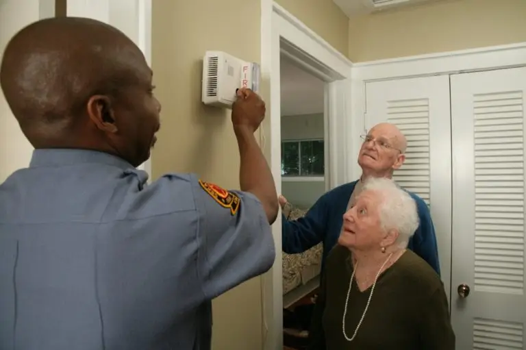 A firefighter is installing a smoke detector in a home while an elderly couple, one of whom has disabilities, watches attentively.