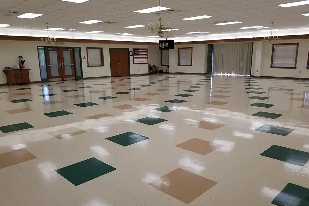 A spacious, vacant room featuring a checkered floor with alternating light and dark tiles, a chandelier hanging from the ceiling, and an american flag displayed next to closed doors.